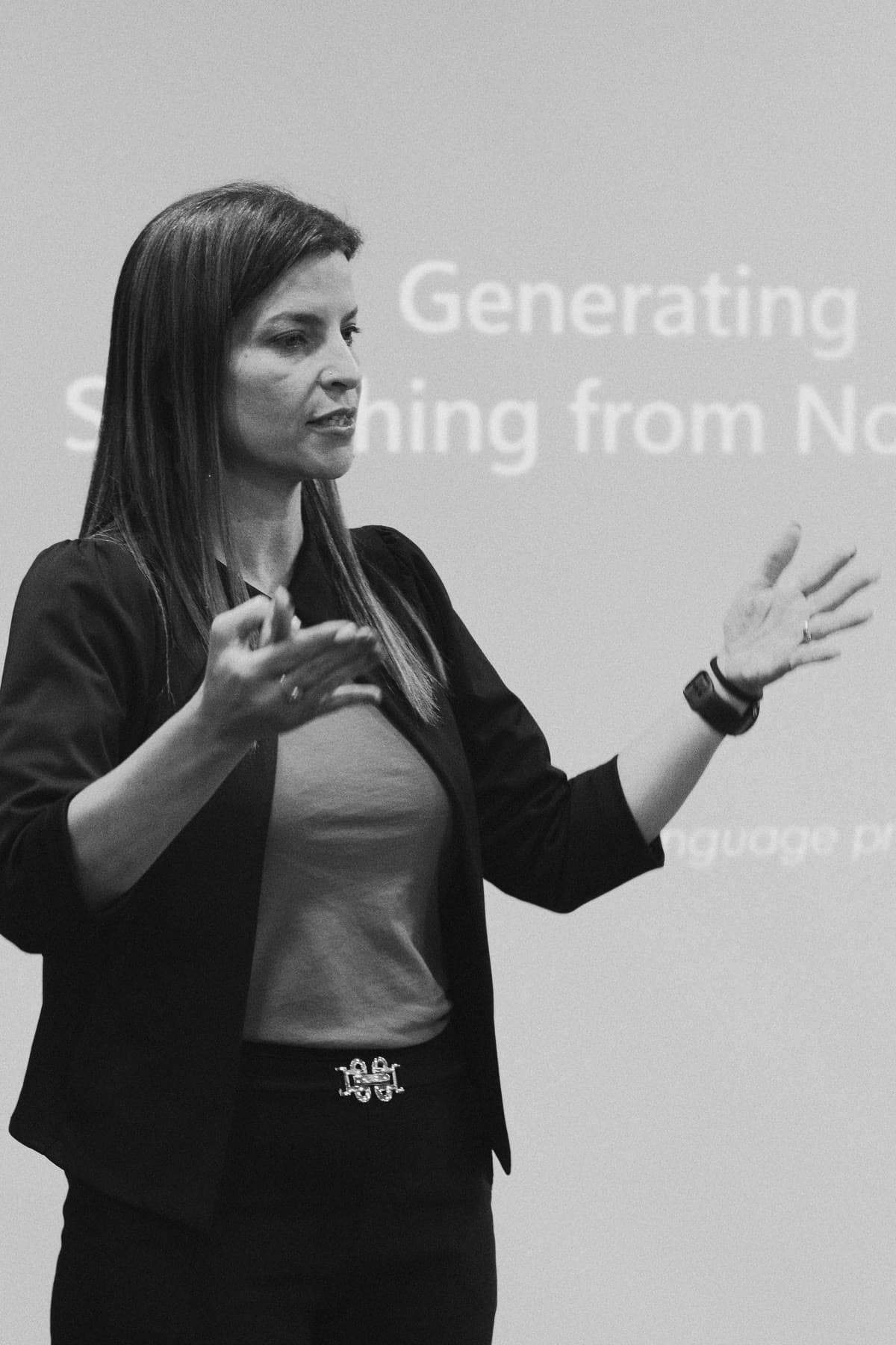 Black-and-white photo of Ady Mor-Biran presenting, both hands raised mid-explanation, her slide behind her.