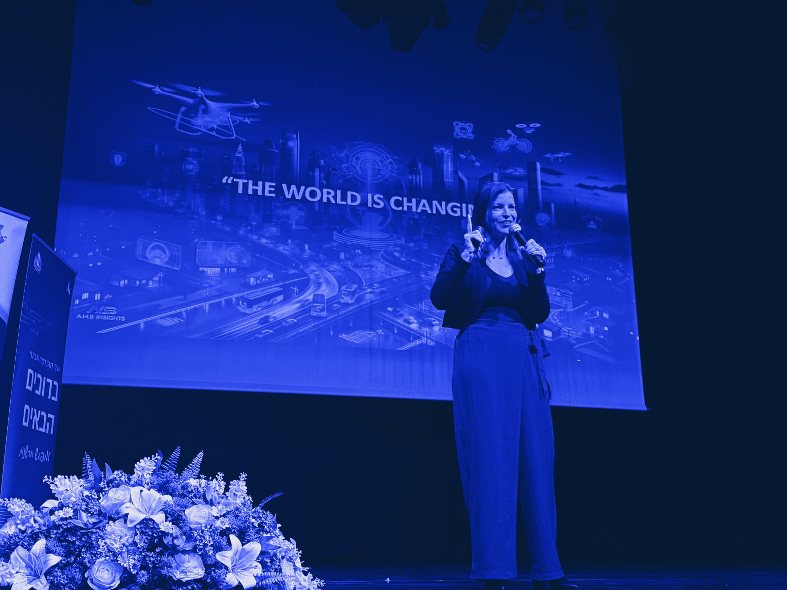 Ady Mor-Biran on a theatre stage, microphone in hand, in front of a slide reading "The world is changing".